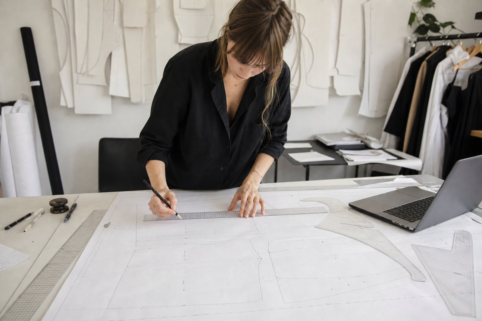 A pattern designer marking a paper sewing pattern with a ruler on a large drafting table beside a laptop